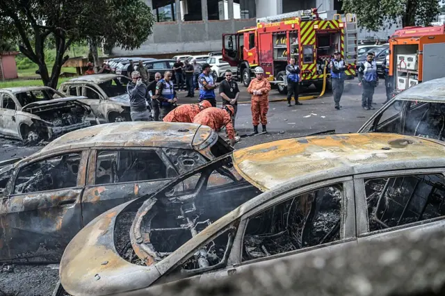 Bombeiros e policiais trabalham ao lado de veículos incendiados no local da queda de helicópteros no bairro do Recreio dos Bandeirantes