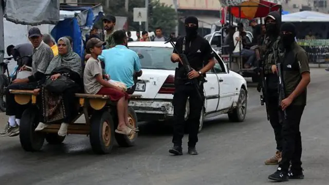 سه نیروی حماس که صورتهایشان را  پوشانده شده، در خیابانی در غزه گشت میزنند. یک ماشین سفید، گاری زرد کوچکی را میکشد که یک زن، چند مرد و یک پسر در آن نشسته و پاهایشان بیرون آویزان است