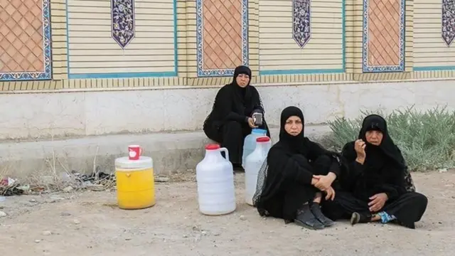 چند زن خوزستانی در حالی که نشسته‌اند، به دوربین نگاه می‌کنند و چند دبه اب کنارشان روی زمین است.