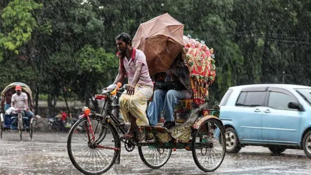 বৃষ্টির মধ্যে একজ চালক রিকশা চালাচ্ছেন, যাত্রী ছাতা ধরে আছে। পেছনে আরেকটি রিকশা ও একটি প্রাইভেটকার দেখা যাচ্ছে। 