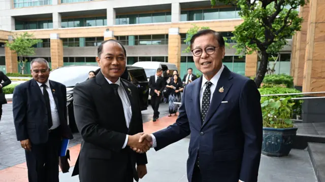 နေပြည်တော်နိုင်ငံခြားရေးဝန်ကြီး ဦးတင်မောင်ဆွေ(ဝဲဘက်)နဲ့ ထိုင်းနိုင်ငံခြားရေးဝန်ကြီး မစ္စတာ ဆီဟာဆက်(ယာဘက်) တို့ ဘန်ကောက်မြို့က ထိုင်းနိုင်ငံခြားရေးဝန်ကြီးဌာနမှာ ဇူလိုင်လ ၁၄ ရက်တုန်း တွေ့ဆုံမှုလို့ဆိုပြီး ထိုင်းဘက်က ထုတ်ပြန်လာတဲ့ဓာတ်ပုံ။ အနောက်တိုင်းဝတ်စုံ ၀တ်ထားတဲ့အမျိုးသား ၂ ဦး ကင်မရာကို ပြုံးပြပြီး လက်ဆွဲနှုတ်ဆက်နေ၊ အနောက်ပိုင်းမှာ အနောက်တိုင်းဝတ်စုံဝတ်ထားတဲ့ အမျိုးသားတစ်ယောက် မတ်တပ်ရပ်နေပြီး၊ ကားအနက်အချို့နဲ့ လူအချို့ကိုလည်း မြင်ရ 
