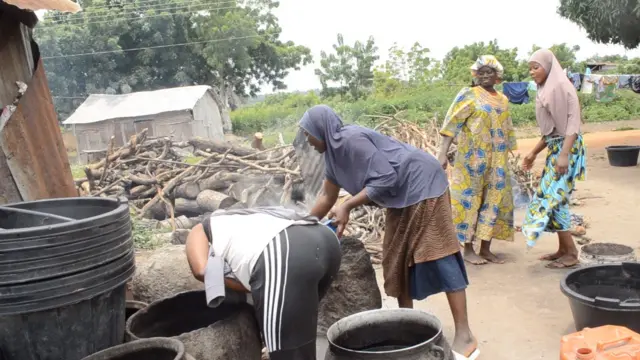Obinrin rin tiwọn wọ oniruuru asọ n se ori. Meji tẹriba lati wo inu ikoko nla dudu ti wn fi n se ori nigba ti awọn obinrin meji duro, igi idana, baafu ipọnmi ati aaro wa ni ayika ibi ti wọn wa.