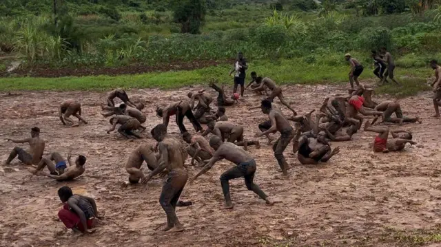 Une trentaine d'hommes s'entraînent à faire des roulades avant sur un terrain boueux en Ouganda.