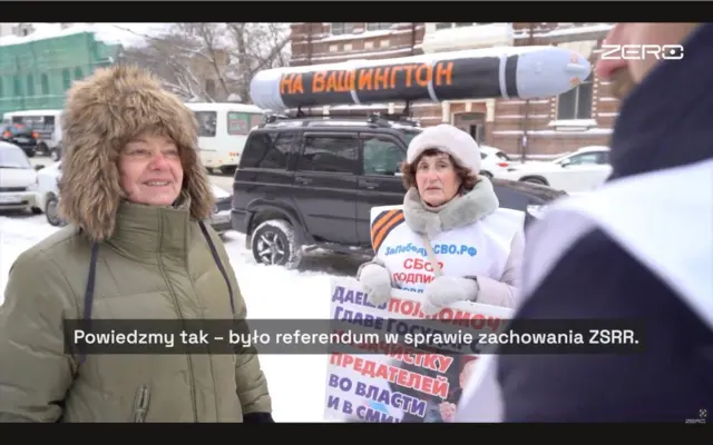 На засніженій вулиці стоять кілька людей, літня жінка у світлому пуховику з хутром тримає плакат із написом російською: "Даєш повноваження голові держави на зачистку зрадників у владі та ЗМІ". Позаду видно автомобіль із муляжем ракети. На передньому плані – Вєрніковська, яка розмовляє з учасником акції. У нижній частині кадру субтитр: "Скажімо так – був референдум щодо збереження СРСР".