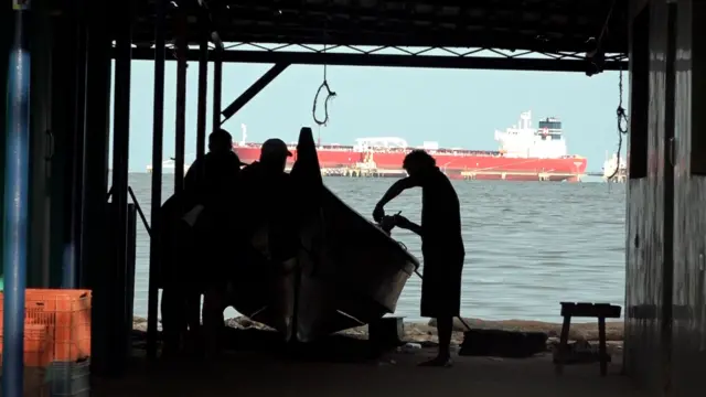 Cuatro hombres a contraluz junto a una lancha en un sitio cubierto frente al mar.