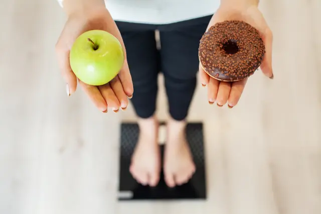 Imagem mostra mulhercáixa loteriaspé sobre balança enquanto segura uma maçã e um donut.