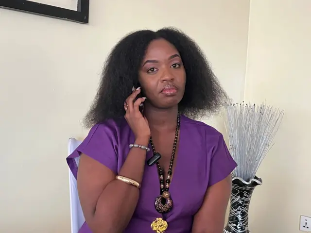 Umugore/umukobwa ufite imisatsi miremire, afite telefone ku gutwi, yambaye umwenda w'iroza w'amaboko magufi, urunigi mu ijosi n'imikufi ibiri ku kuboko, iruhande rwe hari umutako ukoze nk'agacuma urimo amabara y'umukara n'umweru. 