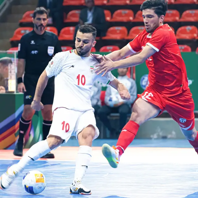 بازیکن شماره ۱۰ ایران با لباس سفید در حال ضربه زدن توپ و بازیکن افغانستان با لباس سرخ تلاش می‌کند که ضربه را دفاع کند.