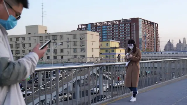Homem e mulher, de máscaras, olham para seus celulares em passarela para pedestres na China