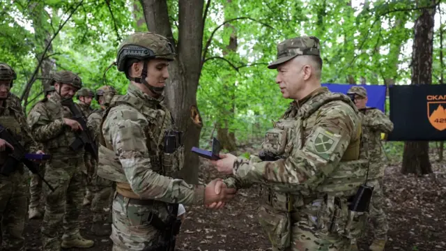Сирський передає бійцю нагороду у лісі, навколо стоять інші військові