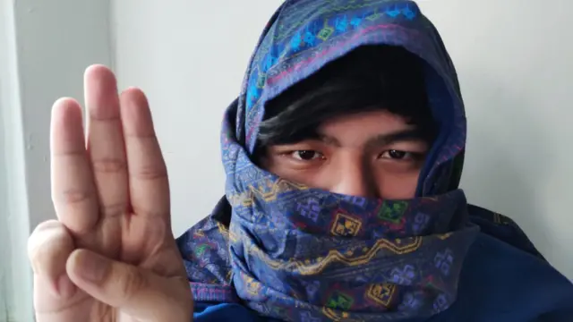 Burmese with purple sarong wrapped round his head doing a three finger salute.