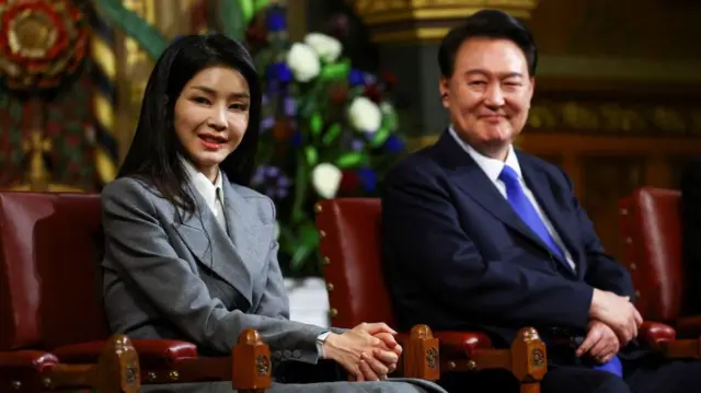 Kim Keon Hee, de terno cinza e sorrindo, sentada em uma cadeira de couro vermelha, ao lado do seu marido, o ex-presidente Yoon Suk Yeol