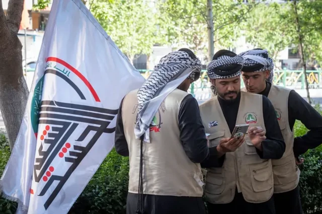 اعضای حشد شعبی عراق در جریان شرکت در تجمعی ضد آمریکا و اسرائیل در مقابل سفارت عراق در مرکز تهران، ۲۳ آوریل ۲۰۲۶
سه مرد با دشداشه و جلیقه‌های کرم رنگ که نشان حشد شعبی روی آنهاست، پشت آنها پرچم سفید رنگی با نشان حشد شعبی