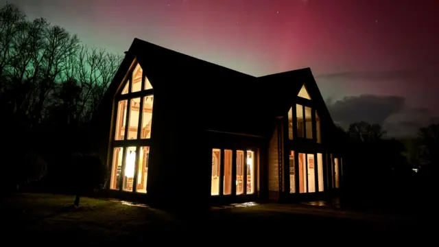 Un edificio bien iluminado en primer plano con el resplandor rojo y verde de la aurora boreal en el fondo, más allá de la línea de árboles.