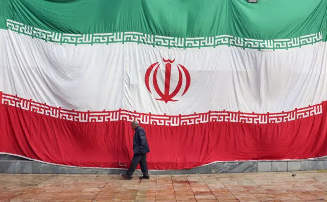 مردی در حال راه رفتن مقابل ساختمانی که در جنگ آسیب دیده و با پرچم بزرگ ایران پوشانده شده است