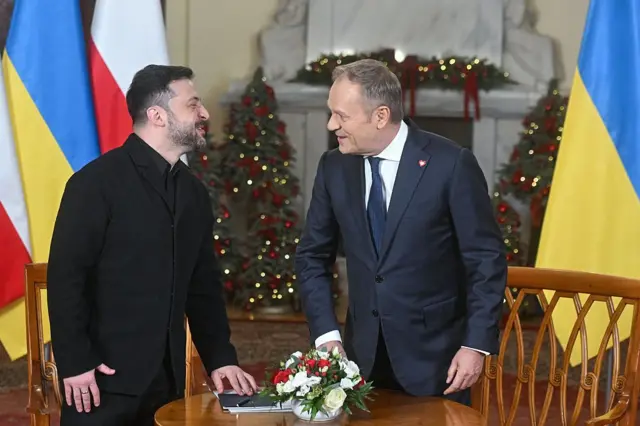 Зеленский (слева) и Туск (справа) садятся за небольшой круглый столик во время официальной встречи. Оба улыбаются, смотря друг на друга. На фоне  —  флаги Украины и Польши, а также украшенные новогодние елочки.