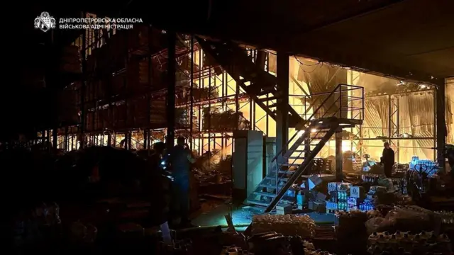 Темне нічне фото зруйнованого складу або виробничої будівлі: всередині яскраво горить світло, видно металеві конструкції й сходи, частково зірваний дах і розкидані речі та ящики з товарами. У приміщенні та зовні стоять кілька людей. 