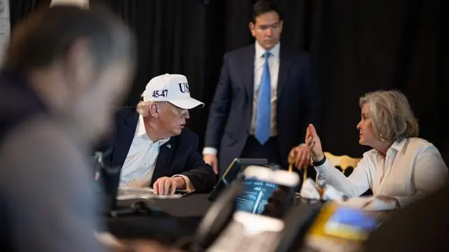 El presidente Donald Trump está sentado con una gorra blanca que dice EE.UU. y habla con la jefa de gabinete de la Casa Blanca, Susie Wiles. En un segundo plano, atrás, está de pie el secretario de Estado, Marco Rubio.