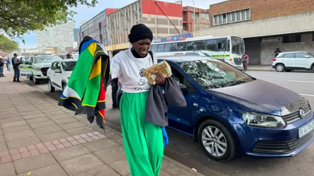 One woman hold South Africa flag and one book. She wear white top and green trouser.