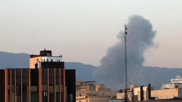 遠く山並みを背に、ベージュ色の複数の建物の合間から黒煙が上っている