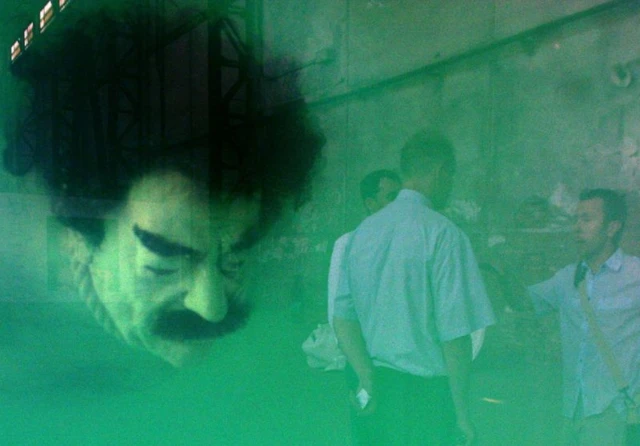 Au premier plan, la tête de la sculpture est plongée dans une eau verte. Au second plan, nous distingons trois hommes discutés entre eux.