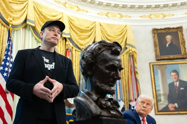 Trump e Musk no Salão Oval da Casa Branca, com quadros a cortinas ao fundo, além da bandeira dos Estados Unidos