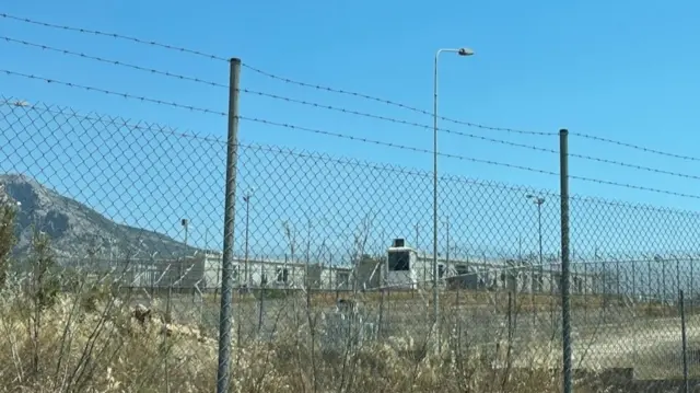 Ogrodzenie z drutu kolczastego, za nim kilka budynków i wzgórze