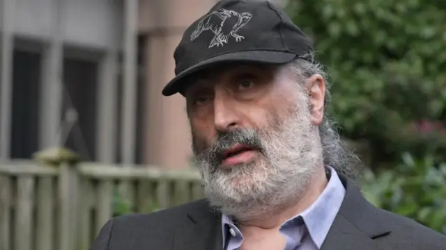 Mark Nygate, un hombre con barba gris, vestido con un traje oscuro y una gorra de béisbol.
