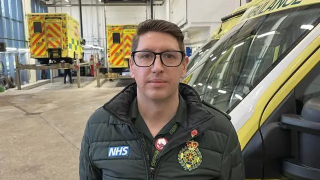 Nakai Redman, gerente de operaciones de SECAMB Ashford, junto a una ambulancia en la zona de preparación. Al fondo, se ven dos ambulancias elevadas con un gato mientras se realizan tareas de mantenimiento.