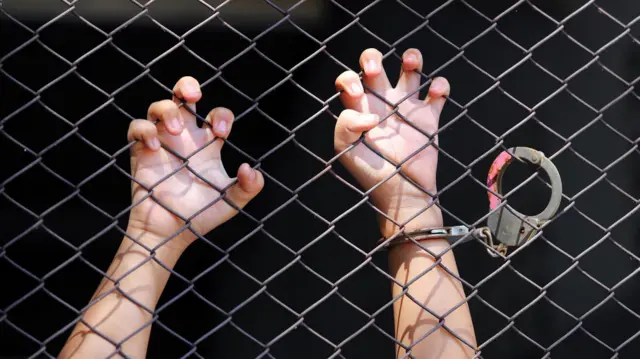 দুটি হাত একটি লোহার গ্রিল খামচে ধরে আছে, একটি হাতে হাতকড়া