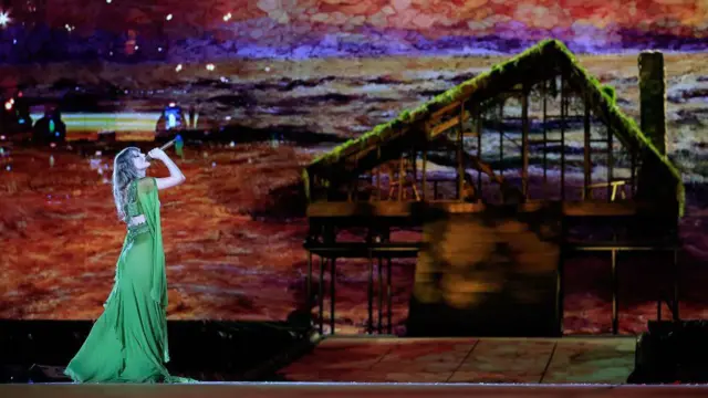 Taylor Swift no palco, usando um vestido longo verde enquanto canta diante de um cenário que reproduz uma cabana de madeira coberta de vegetação. Ao fundo, um grande painel exibe uma paisagem em tons alaranjados e arroxeados.