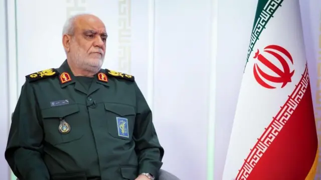 مجید خادمی با لباس نظامی روی صندلی نشسته و در کنار او پرچم جمهوری اسلامی دیده می‌شود