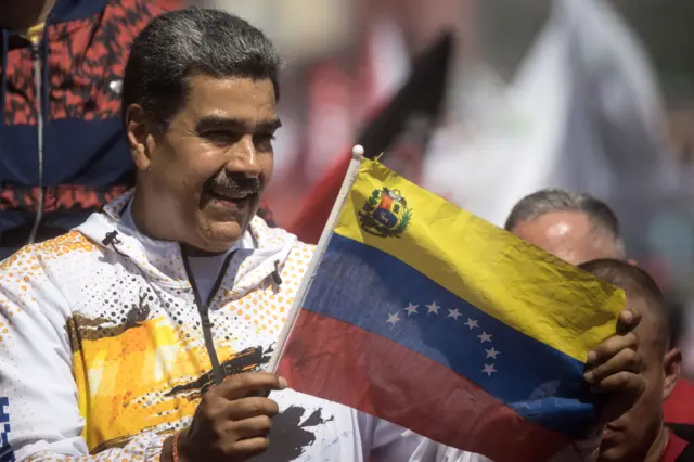 Nicolas Maduro vestindo casaco branco segura bandeira da Venezuelafrutinha caça níquelcomício