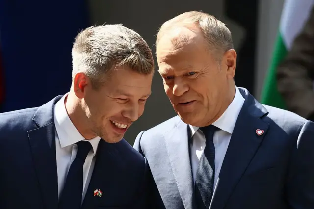 Мадяр і Туск крупним планом стоять поруч на відкритому повітрі. Обидва нахилилися один до одного, спілкуючись. На костюмі Мадяра помітний значок із прапорами Польщі та Угорщини, а в Туска – невеликий значок у формі серця (символ його партії).