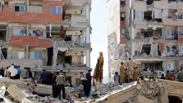 Iran people and soldiers stand around buildings wey don damage
