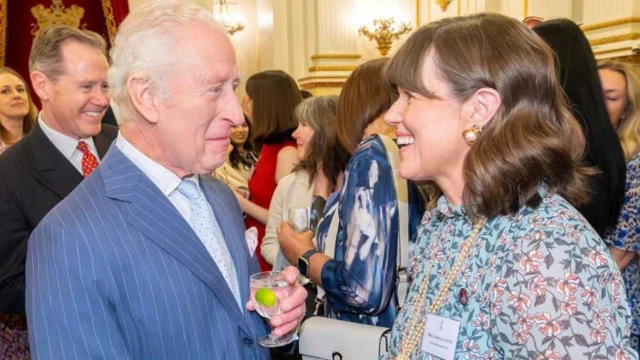 King Charles on the left is wearing a blue suit with a thin white stripe, a white shirt and pale blue tie. He is holding a glass in which there is clear liquid and a slice of lime. He is looking at Caroline Jones and smiling with a closed mouth. Caroline on the right is smiling, she wears a light green floral dress with a long string of pearls and pearl earrings in a gold setting. 