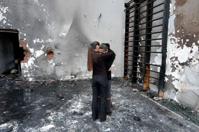 Duas mulheres se abraçando dentro do ginásio destruído, dias após o ataque.