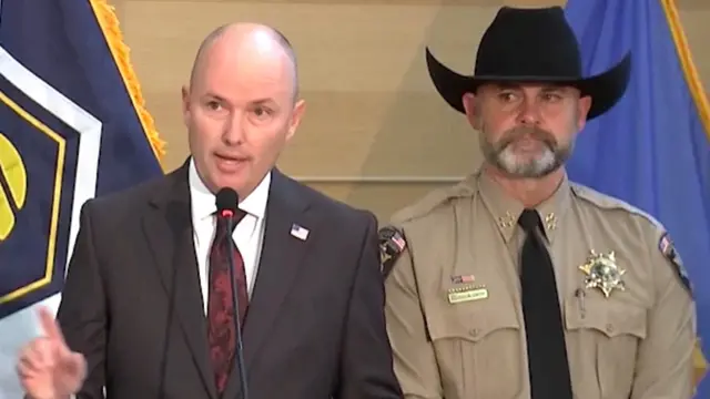 Le gouverneur de l'Utah, Spencer Cox, parle dans un micro, vêtu d'un costume gris et d'une cravate rouge foncé. À ses côtés se tient le shérif du comté de l'Utah, Mike Smith, coiffé d'un chapeau à large bord et vêtu d'une chemise d'uniforme marron avec un insigne de police sur la poitrine gauche et des écussons « Sherif » sur les deux épaules.