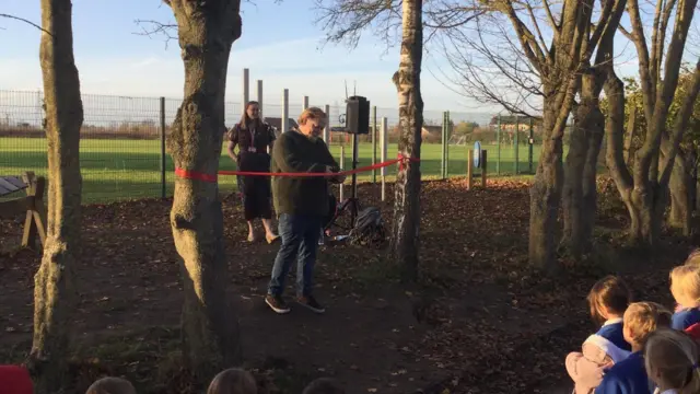 La imagen muestra una escena al aire libre durante un evento de inauguración en una zona boscosa junto a un campo cercado.