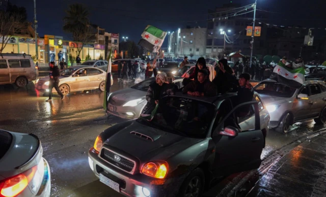 Suriye bayrakları taşıyan bir araç trafikte ve karanlıkta ileriliyor. Neredeyse araçtaki herkes kafalarını camdan çıkarmış durumda. Arkada başk araçlar da var.