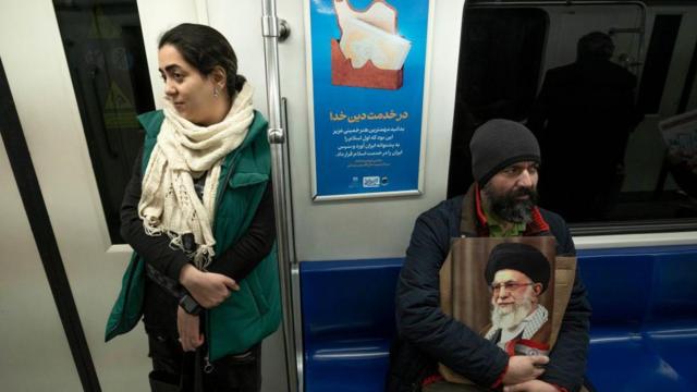 زنی بدون حجاب در مترو ایستاده است و مردی نشسته و عکس آیت‌الله خامنه‌ای را در دست دارد