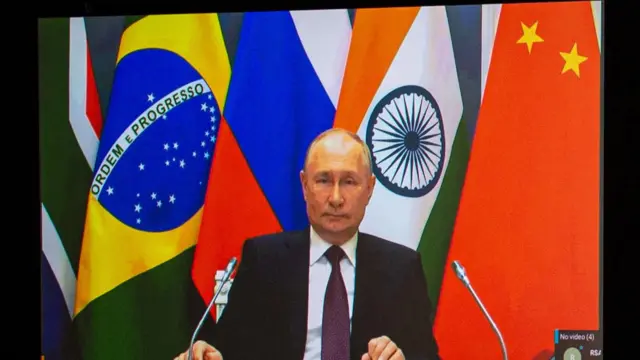 President Vladimir Putin sits as he virtually addresses audience at the Bric summit. Behind is the backdrop of the flags of Brics nations.