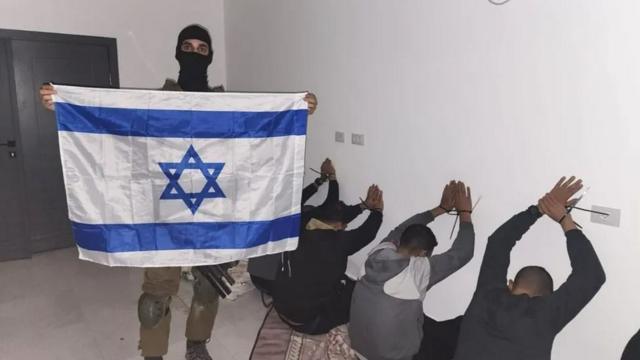 Homem mascarado com bandeira de Israel diante de homens ajoelhados