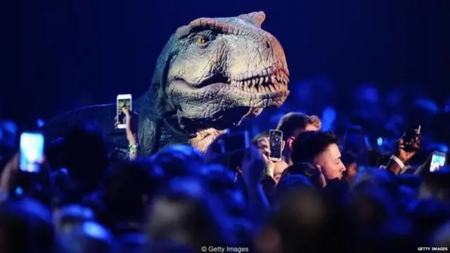 डायनासोरच्या प्रतिकृतीसोबत उभारलेल्या लोकांचा फोटो