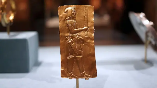 Una placa dorada en la que se ve un hombre de perfil llevando un objeto largo en la mano. Viste un gorro, una túnica corta y pantalones. Lleva una especie de espada en la cintura.
