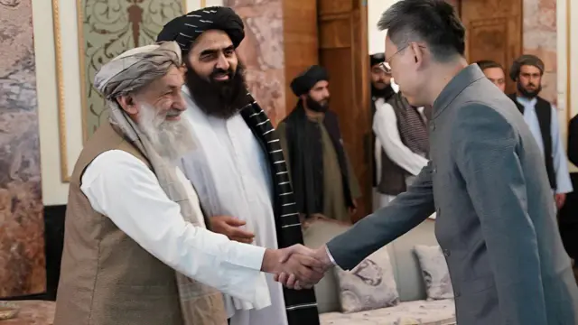 راست سفیر چین با لباس سرمه‌ای که عینک بر چشم دارد با رئیس‌الوزرای حکومت طالبان که لباس سفید، واسکت و لنگی روشن و لبخند دارد دست داده‌است. وزیر خارجه طالبان و چند مقام دیگر در عکس هم دیده می‌شوند.