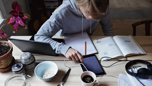 Un joven estudiando sentado. En la mesa tiene un laptop, una tablet, libros y cuadernos de apuntes abiertos y una taza de café