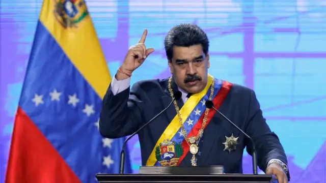 Nicolás Maduro, um homem venezuelano com bigode e cabelos pretos curtos o dedo estirado,terno,frente à bandeira da Venezuela