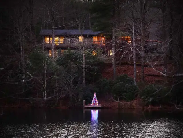 Una casa de dos plantas frente al lago, rodeada de altos árboles, con un árbol de Navidad iluminado. La luz cálida sale a través de las ventanas y las luces del árbol se reflejan en la superficie del lago.