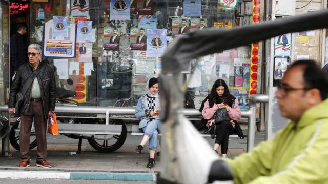 زنی بدون حجاب در کنار زنی با روسری عقب رفته در ایستگاه اتوبوسی نشسته‌اند و مردی در کنار آنها ایستاده است و تبلیغات انتخاباتی پشت آنها دیده می‌شود